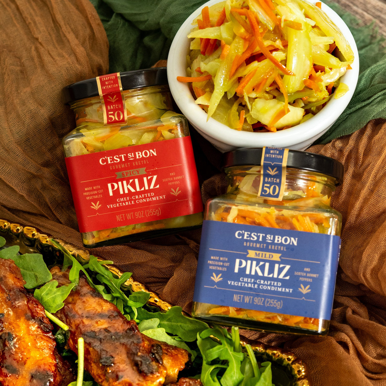 C’est Si Bon mild and spicy pikliz jars beside a bowl of pikliz and a plated meal