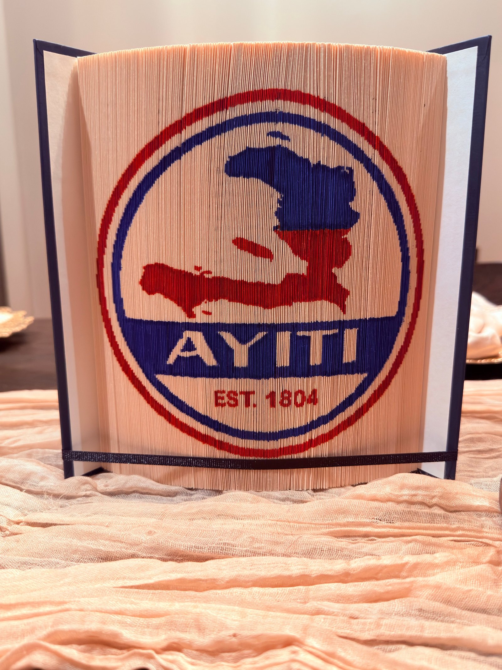 Folded-book artwork with a red and blue Haiti outline and the words AYITI, EST. 1804
